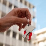 Mână care ține cheile unei locuințe, cu clădiri rezidențiale în fundal, simbol pentru tranzacții imobiliare și servicii de cadastru e-Terra.