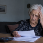 Pensionară în vârstă, îngrijorată în timp ce citește facturi și documente bancare la masa din locuința sa.